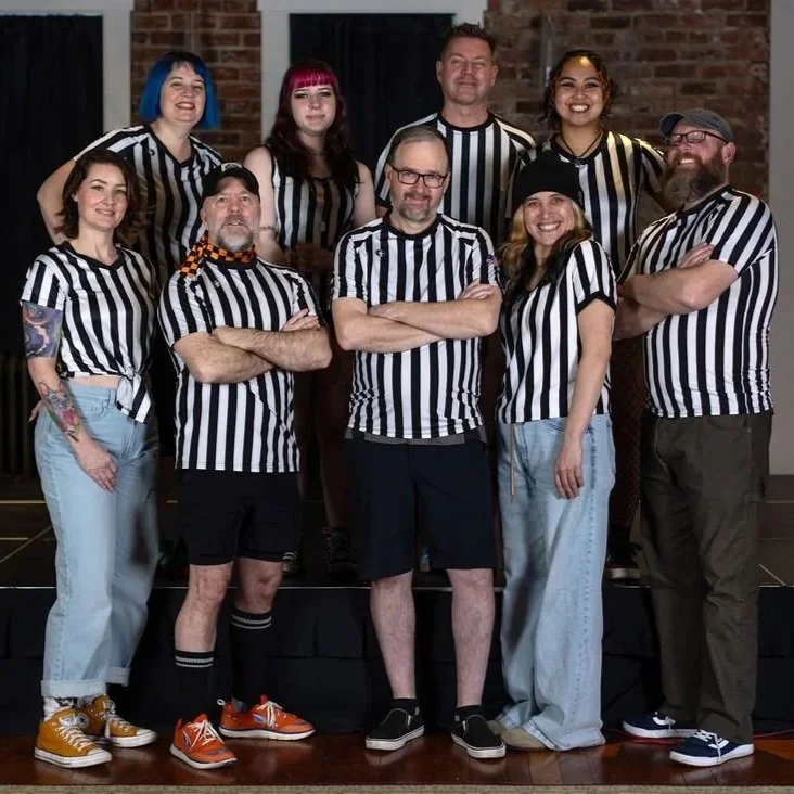 A group of roller derby officials in striped referee shirts pose together in front of a logo that reads 'Tomorrowland Junior,' on a wooden floor.