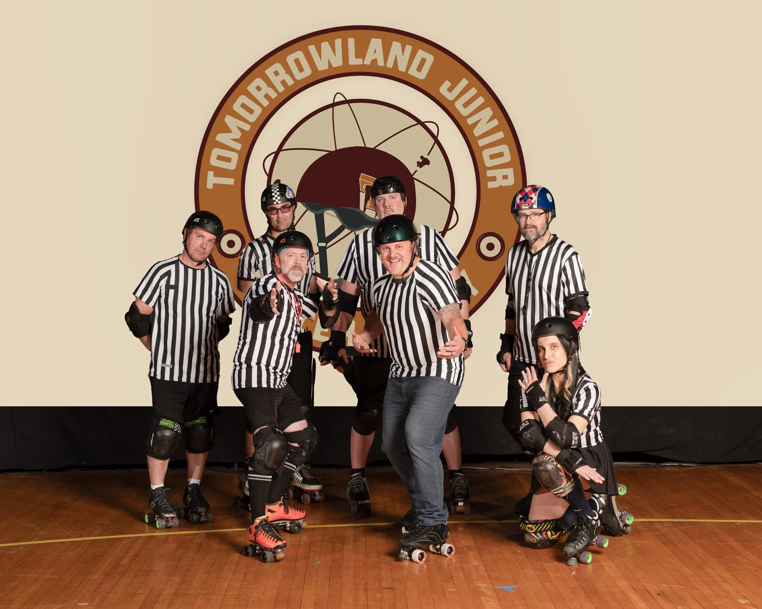 A group of roller derby officials in striped referee shirts pose together in front of a logo that reads 'Tomorrowland Junior,' on a wooden floor.
