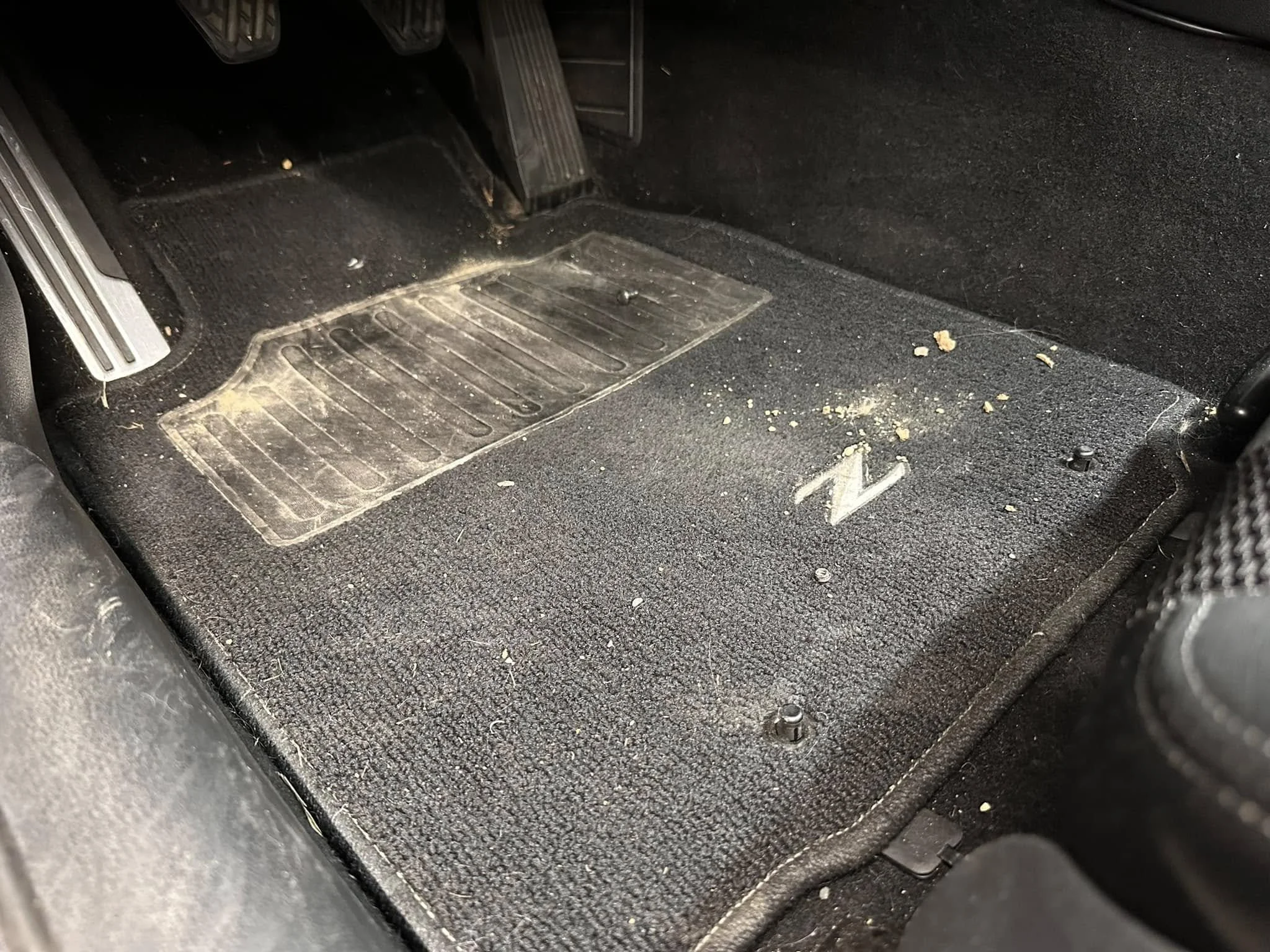 Car floor mat with dirt, crumbs, and debris on black carpet, next to brake pedal and accelerator pedal.
