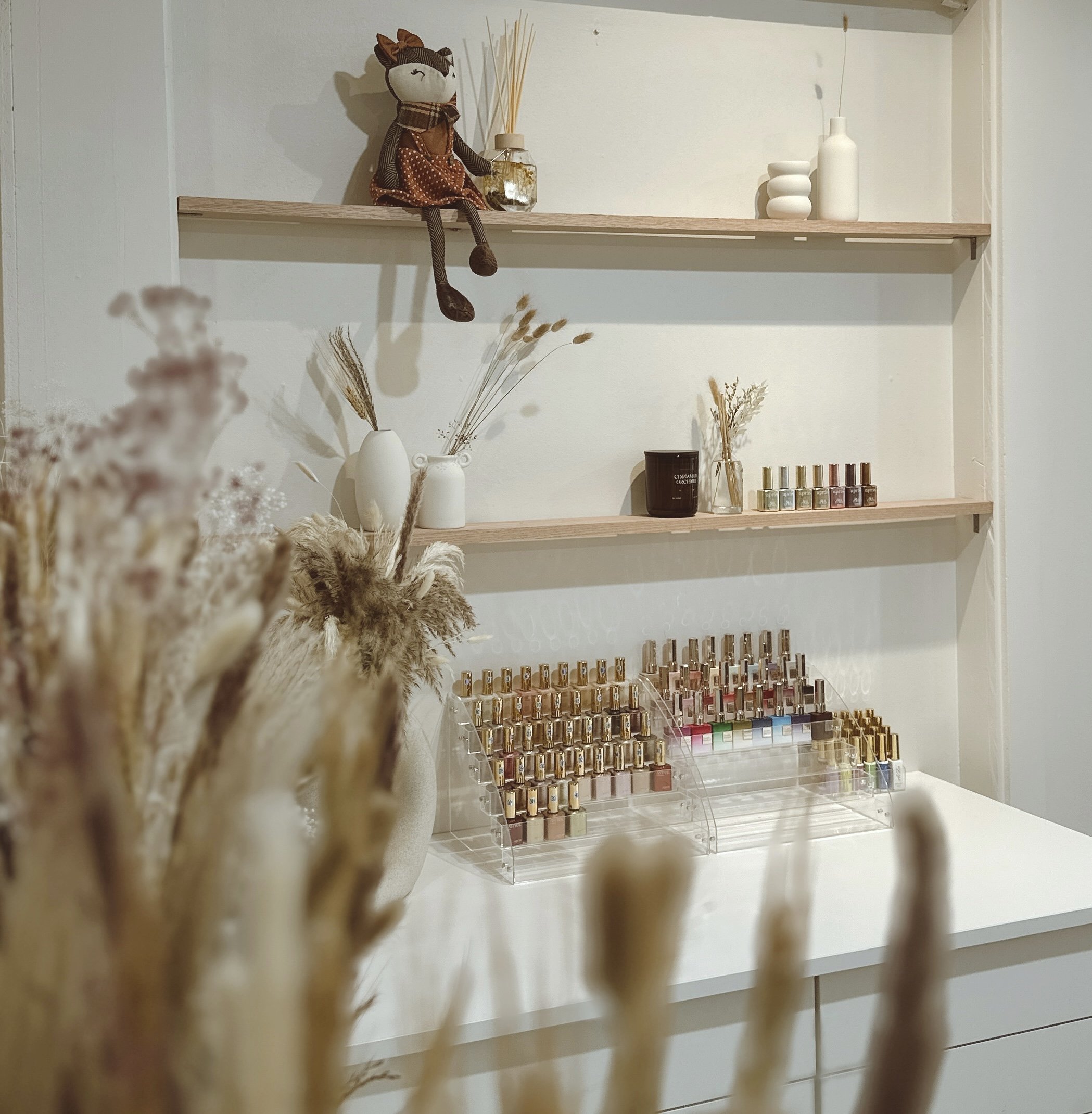 A display of nail polish bottles arranged in clear acrylic stands on a white surface with dried flower arrangements and minimalist decorations on wooden shelves against a white wall.