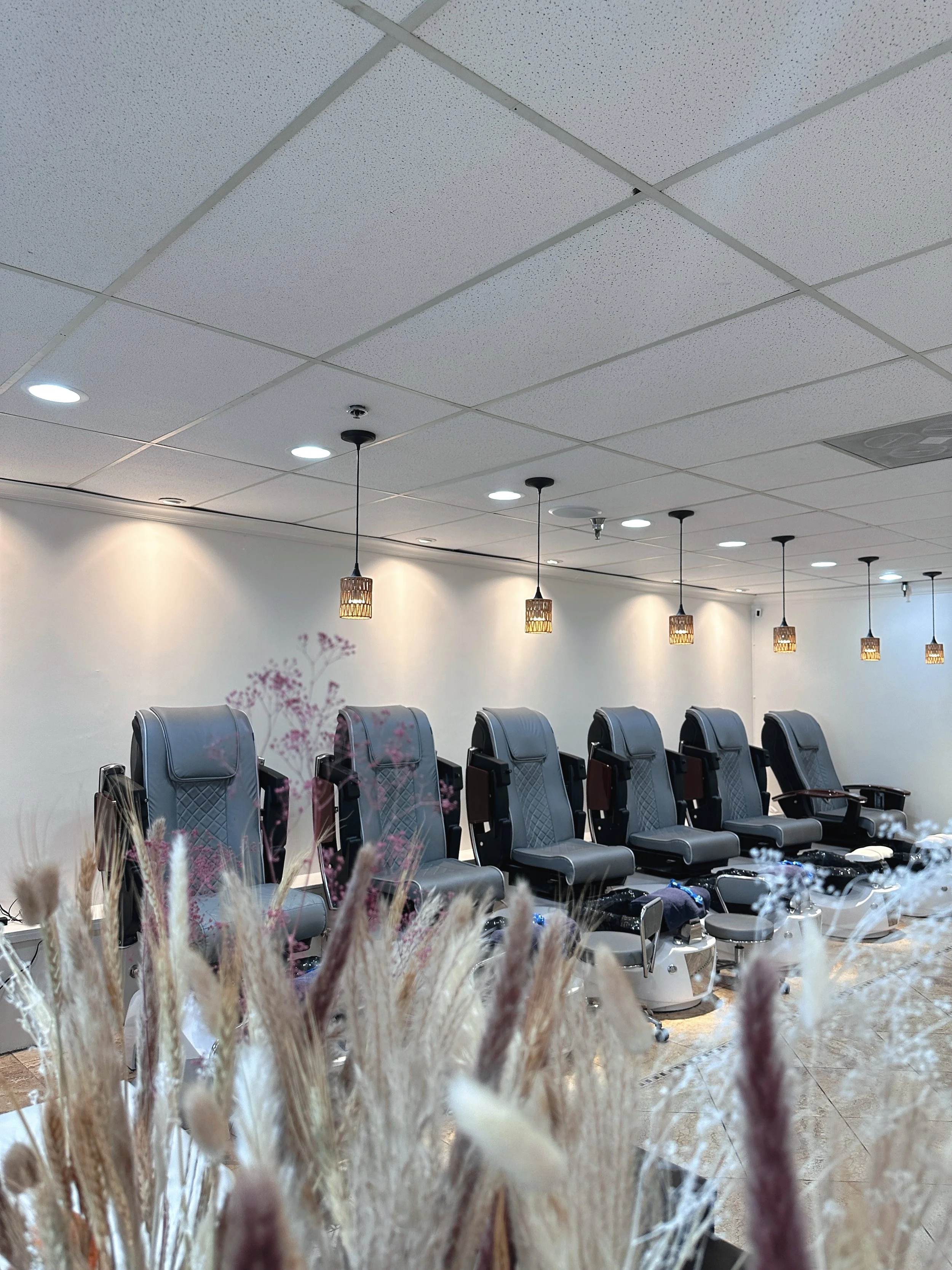 A row of six grey massage chairs in a wellness or spa facility, with hanging pendant lights and a plain wall in the background, and dried decorative plants in the foreground.