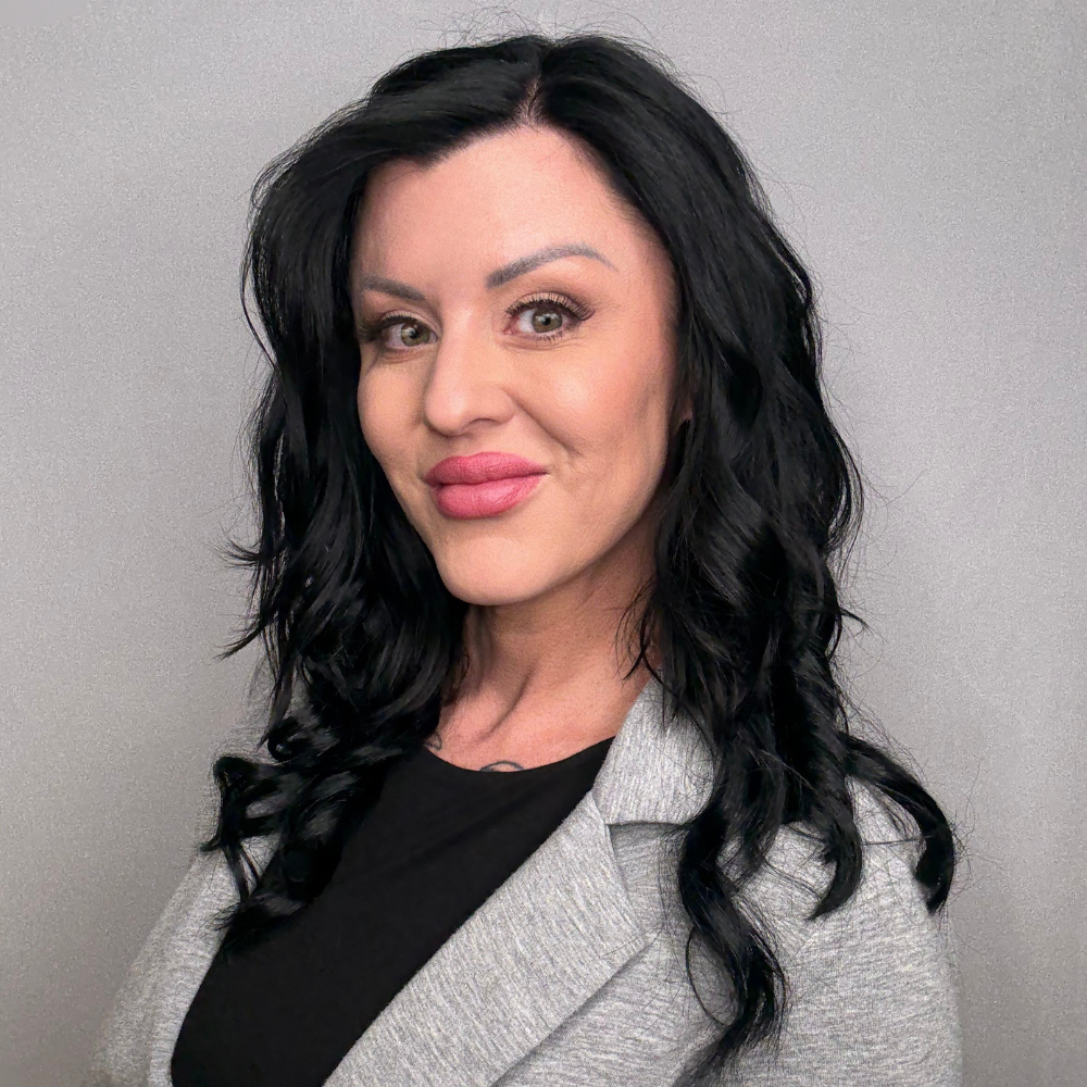 A woman with black, wavy hair, wearing a grey blazer over a black top, posing against a plain grey wall.