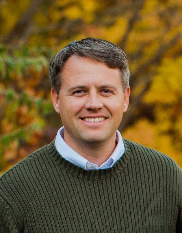 A man smiling outdoors with autumn trees in the background.