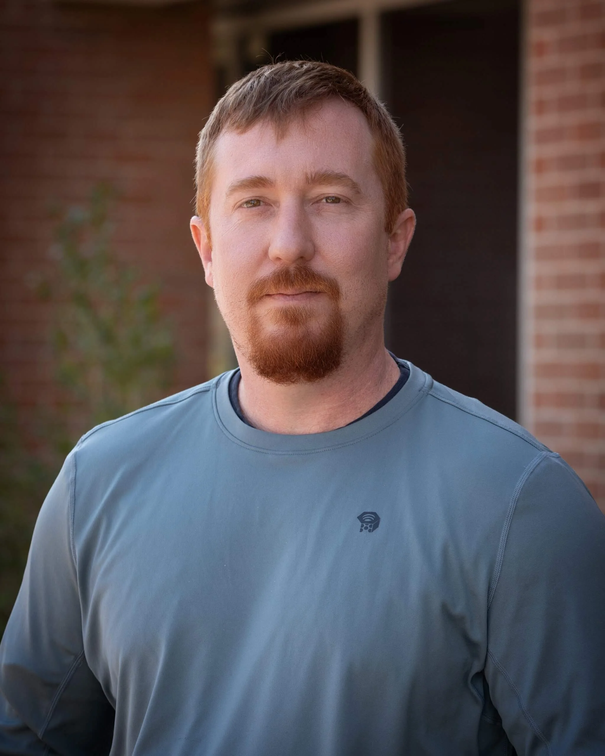 A young man with red hair, beard, and mustache wearing a light blue athletic shirt outdoors with a brick building in the background.