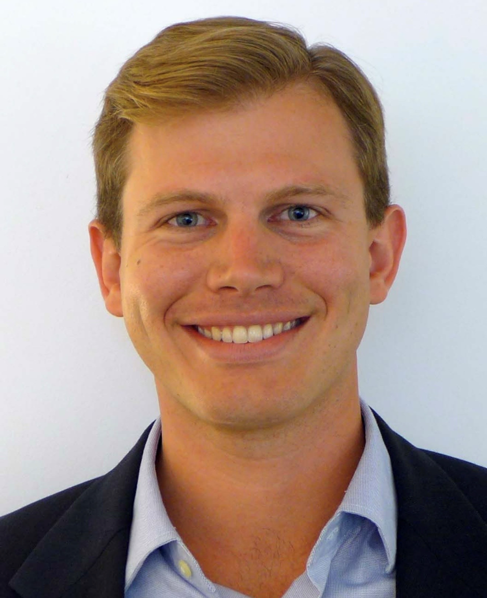 A young man with short glasses and blue eyes, smiling, in professional attire against a white background.