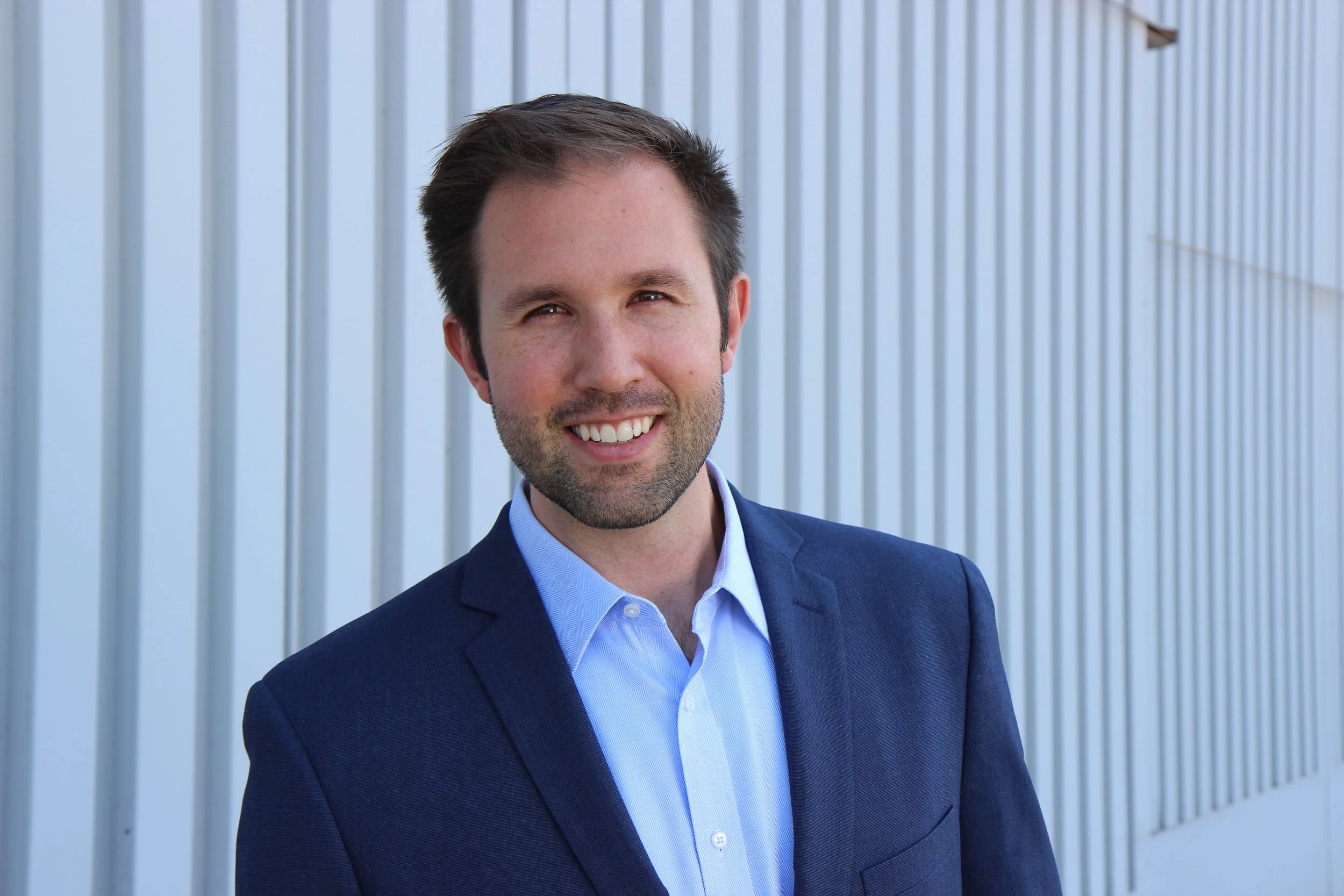 Man in a navy blazer and light blue shirt smiling, standing outdoors in front of a white metal panel wall.