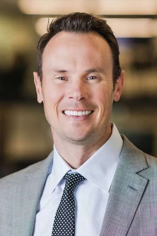 A smiling man wearing a light gray suit, white shirt, and black polka dot tie, with short neatly styled dark hair, in an indoor setting.