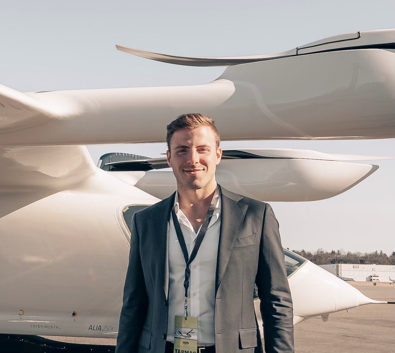 A man in a gray suit and white shirt standing outdoors in front of futuristic white aircraft or drones at an airfield.