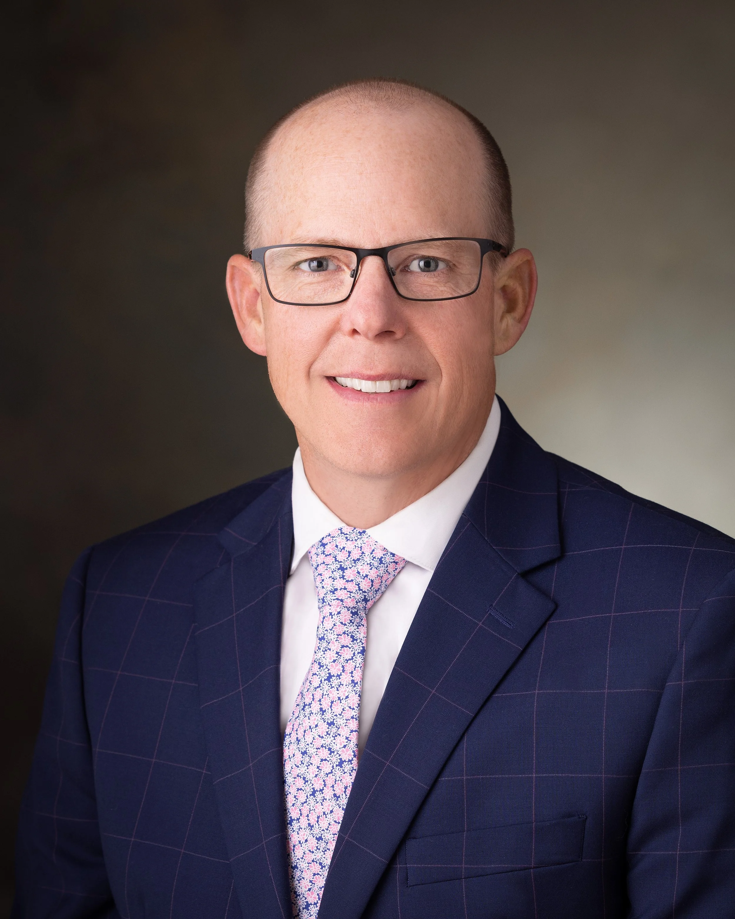 Headshot of a man with glasses, wearing a suit and a light-colored tie with a floral pattern, smiling against a neutral background.