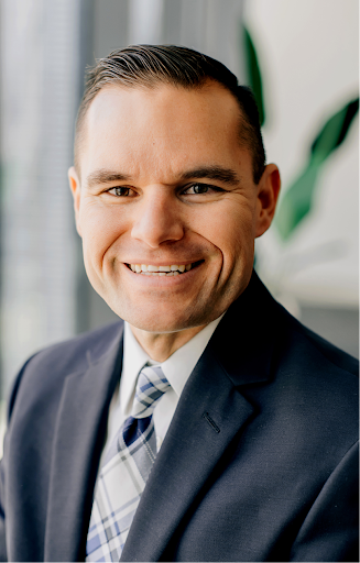 A smiling man in a business suit with a blue plaid tie, indoors with a blurred background including green plants.