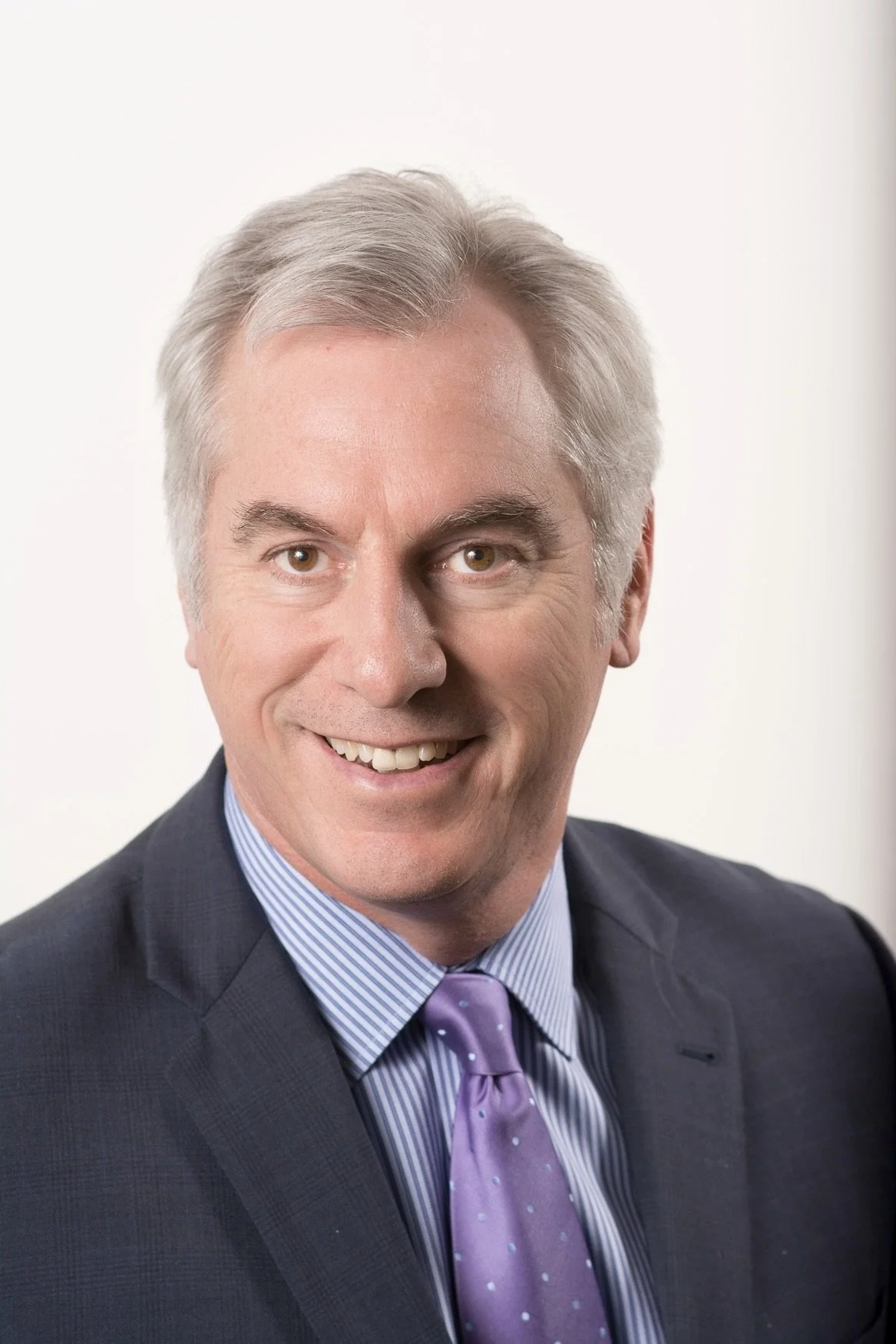 Headshot of a middle-aged man with gray hair, wearing a dark professional suit, blue striped shirt, and lavender tie, smiling against a plain light background.