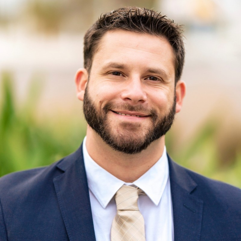 A smiling man with a beard wearing a dark blue suit, white shirt, and beige tie outdoors with blurred greenery in the background.