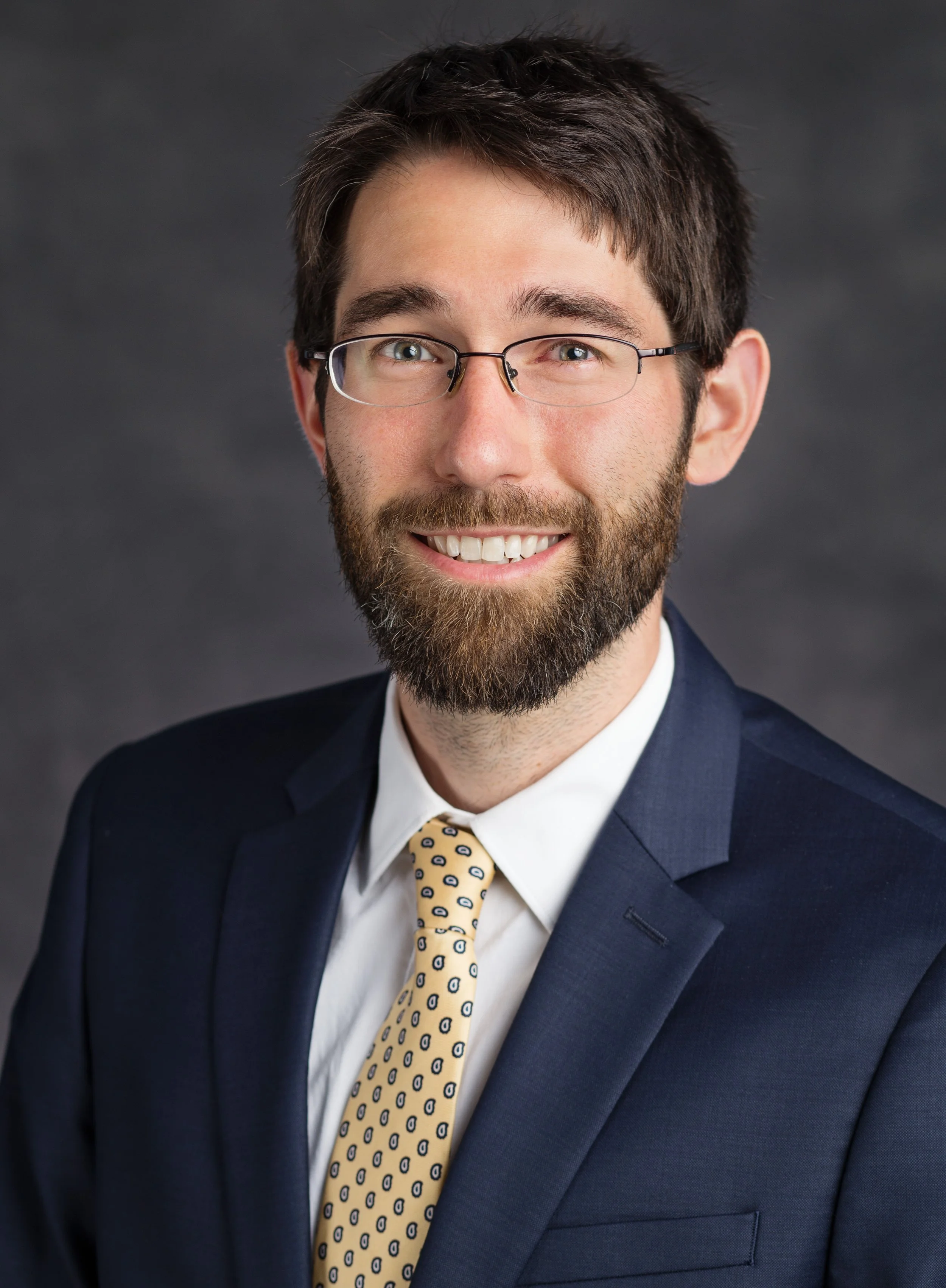 A man with dark brown hair, a full beard, and glasses, wearing a navy blue suit, white shirt, and a yellow tie with blue circles, smiling against a dark gray background.