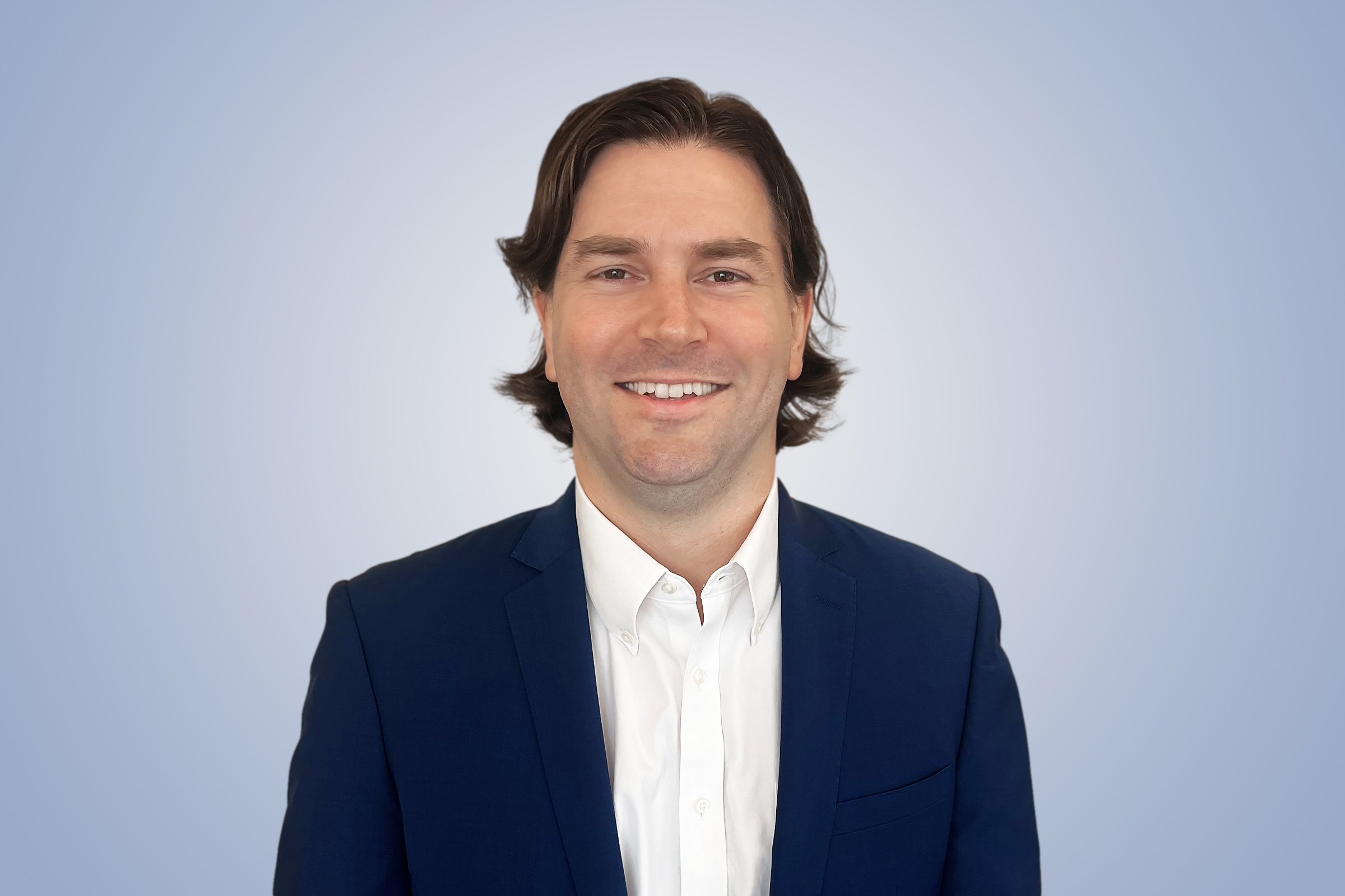 Headshot of a smiling man with brown hair, wearing a dark blue suit jacket and white shirt, against a light blue background.