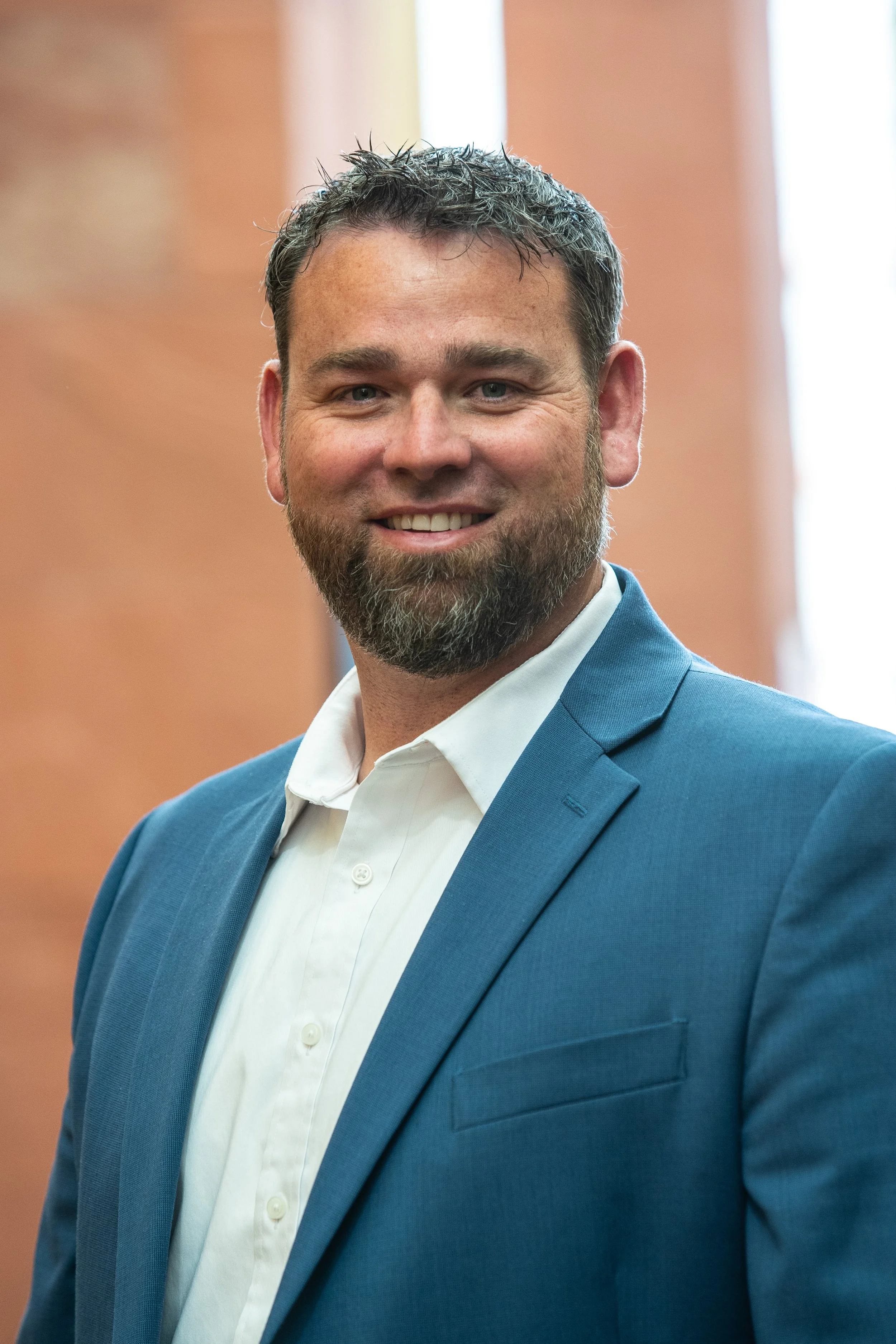 A smiling man with a beard in a blue suit and white shirt, indoor setting with blurred wooden wall and window in the background.