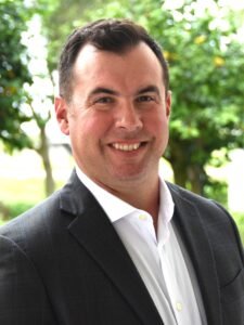 Portrait of a man in a black suit and white shirt, outdoors with green trees in the background.