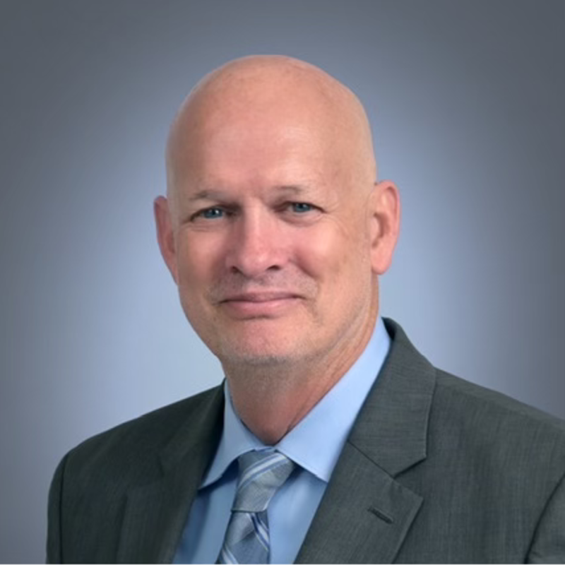 Professional portrait of a bald man in a gray suit and light blue shirt with a tie, smiling slightly against a gray background.