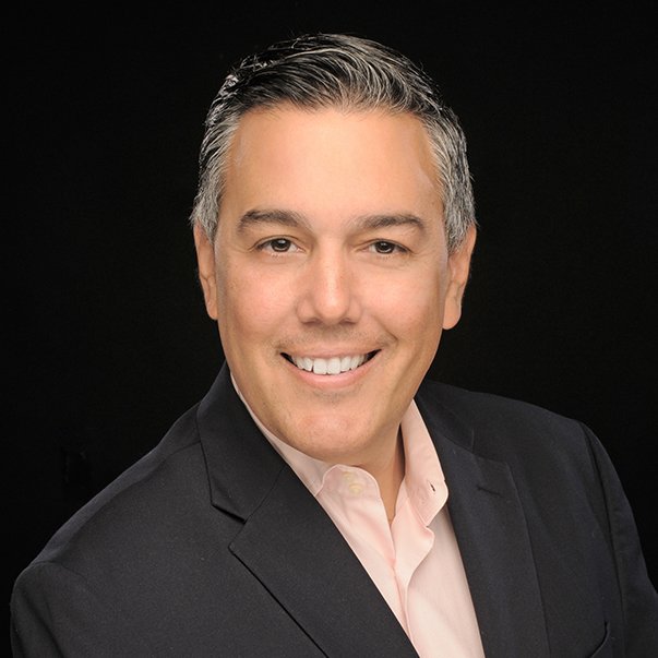 Professional portrait of a smiling man in a dark suit and light pink shirt against a black background.