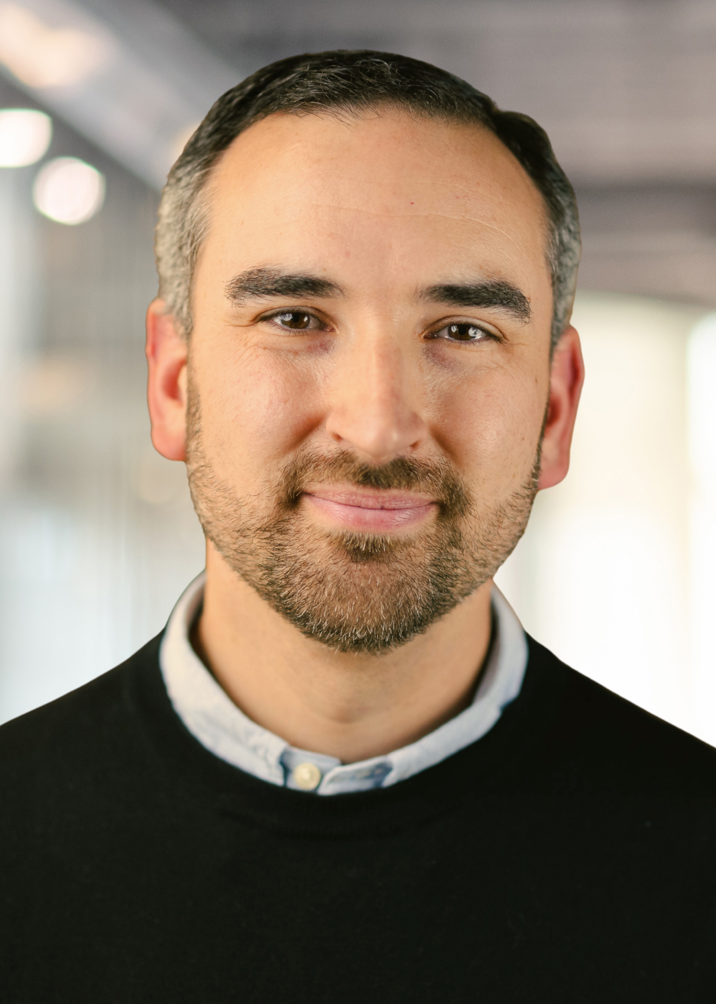 Close-up of a smiling man with short dark hair, light facial hair, wearing a black sweater over a collared shirt, standing in a blurred indoor setting with bright lighting.