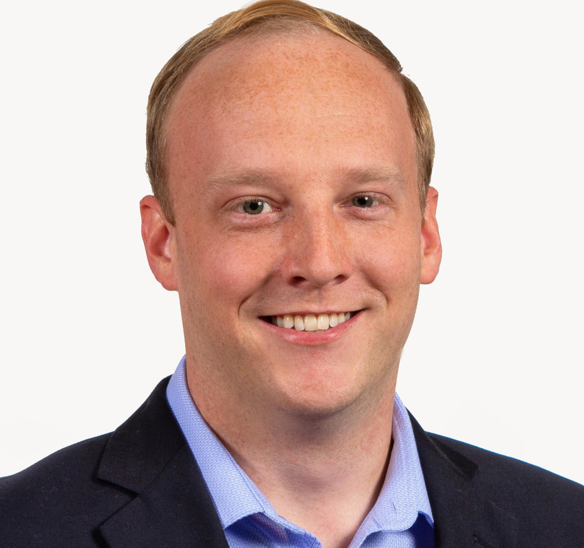 A portrait of a smiling man with short blonde hair wearing a dark suit and light blue shirt against a plain white background.