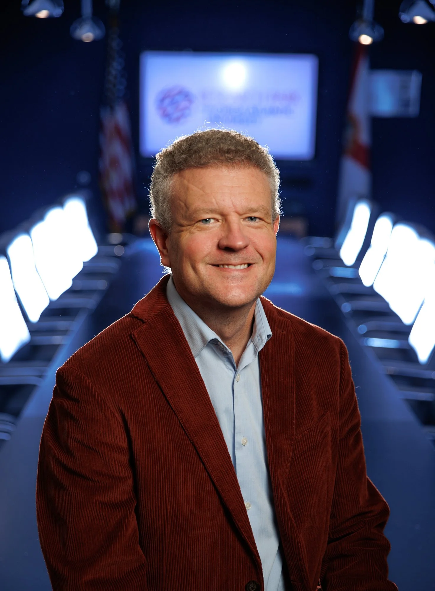 A man in a red blazer and light blue shirt smiling in front of a blue-lit conference room with American flags.
