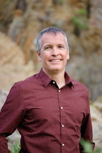 A smiling man with short gray hair wearing a maroon button-up shirt outdoors with rocks and greenery in the background.