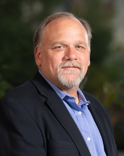 A middle-aged man with a beard and mustache, wearing a dark blazer over a blue shirt, standing outdoors with greenery in the background.