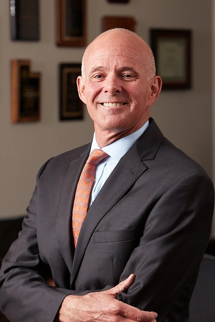 A smiling, bald man in a dark suit, light blue shirt, and patterned orange tie standing with arms crossed in an office setting with framed certificates or awards on the wall behind him.