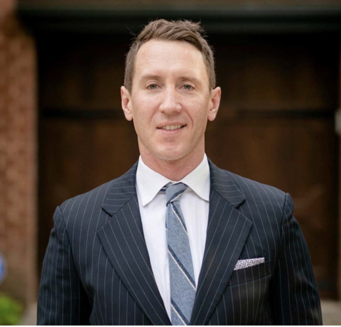 A man with short brown hair, wearing a dark pinstripe suit, white shirt, and a light-colored striped tie, standing outdoors in front of a brick building with a dark background.