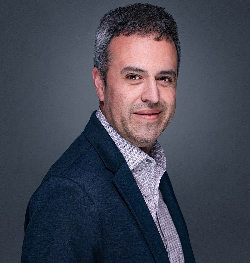 A headshot of a man with short, dark hair, wearing a dark blazer and light dress shirt, smiling slightly against a gray background.