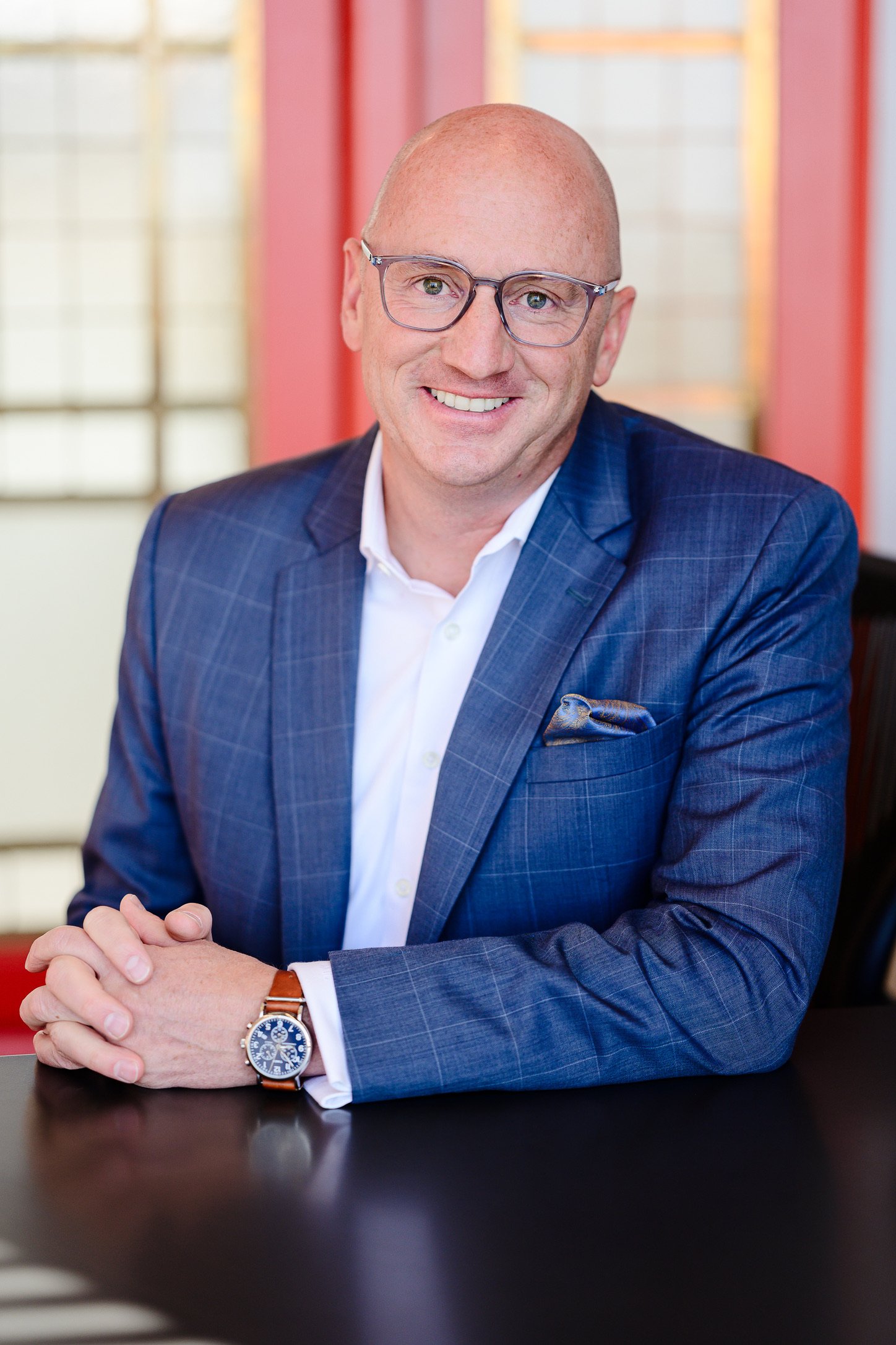 Man in a blue suit jacket and white shirt, wearing glasses and a watch, sitting at a table with clasped hands, in an indoor setting with blurred red and cream-colored background.