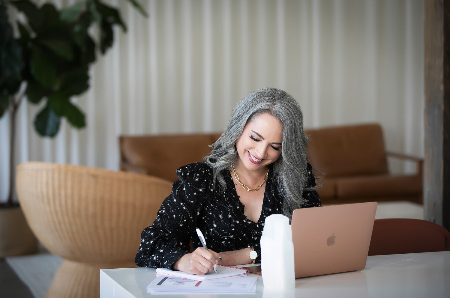 A woman with gray hair working at a white desk with a laptop, notebook, pen, and tissue, smiling and writing.