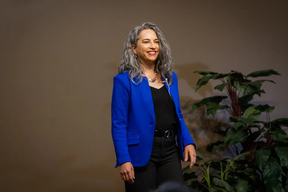 A woman with gray, curly hair is standing against a plain background, wearing a bright blue blazer, black top, and black pants. She is smiling and looking slightly to her right.