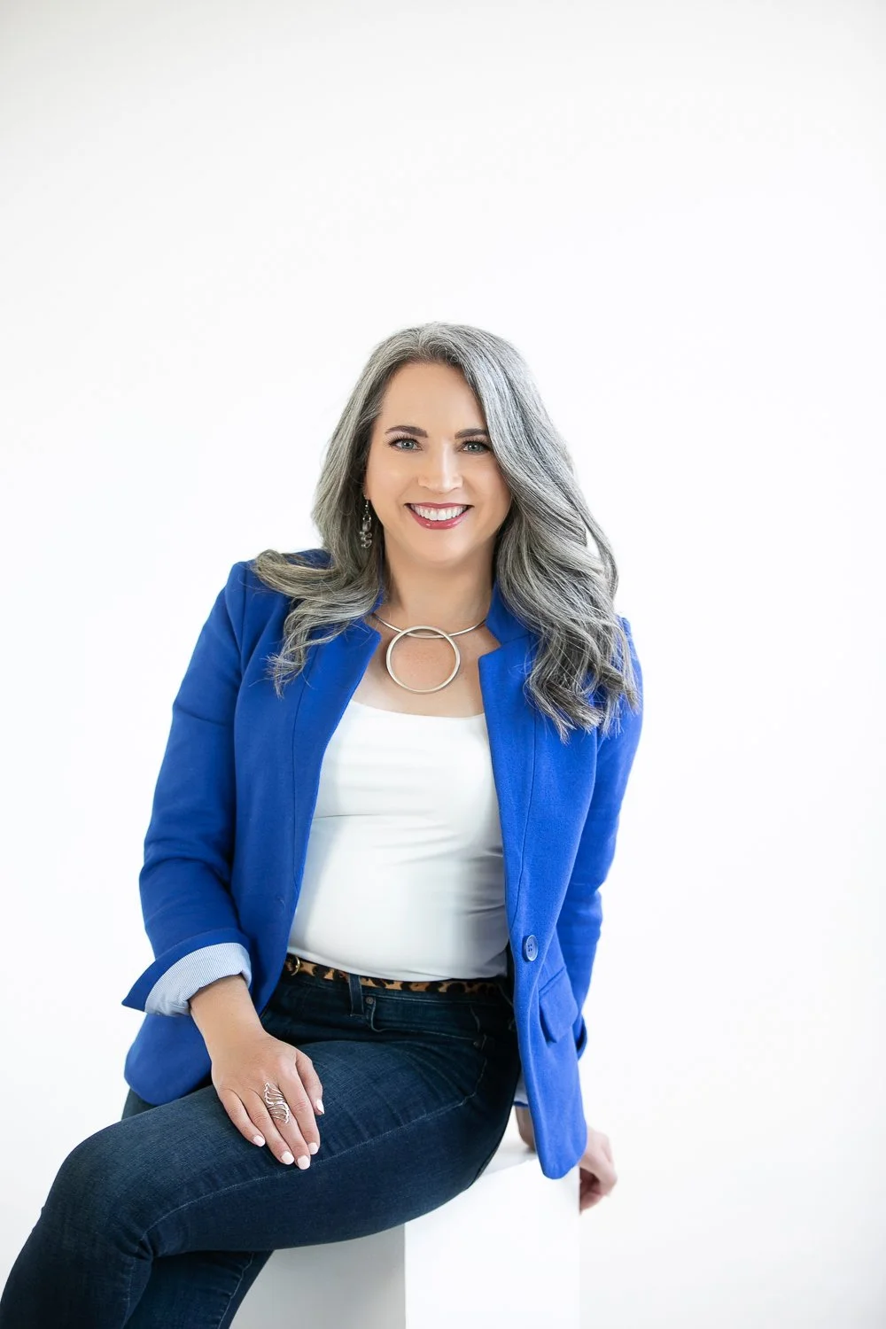 A smiling woman with gray hair wearing a blue blazer, white top, and dark jeans, sitting on a white surface against a plain white background.