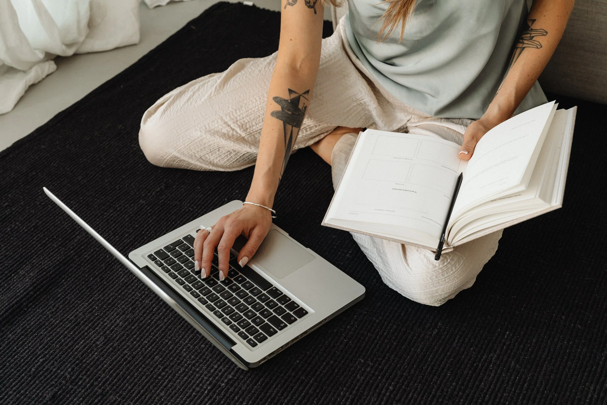A person sitting cross-legged on a black carpet, using a laptop with their left hand, and holding an open planner with a pen in their right hand. They are wearing light-colored pants and a loose, pale-colored top, with visible tattoos on their arms.