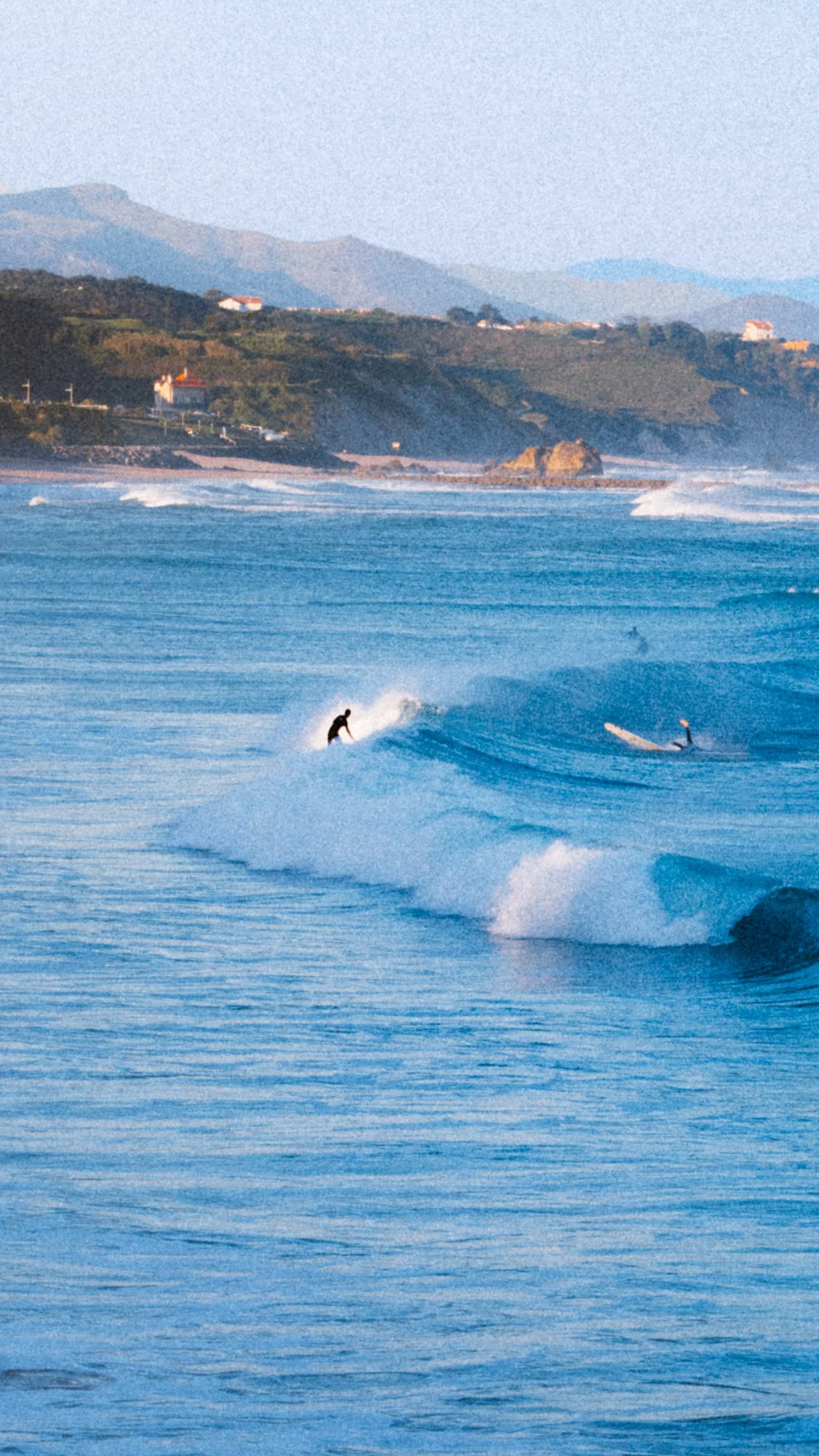 Two surfers riding waves in the ocean with a coastal landscape and mountains in the background during the daytime. Basque coast.