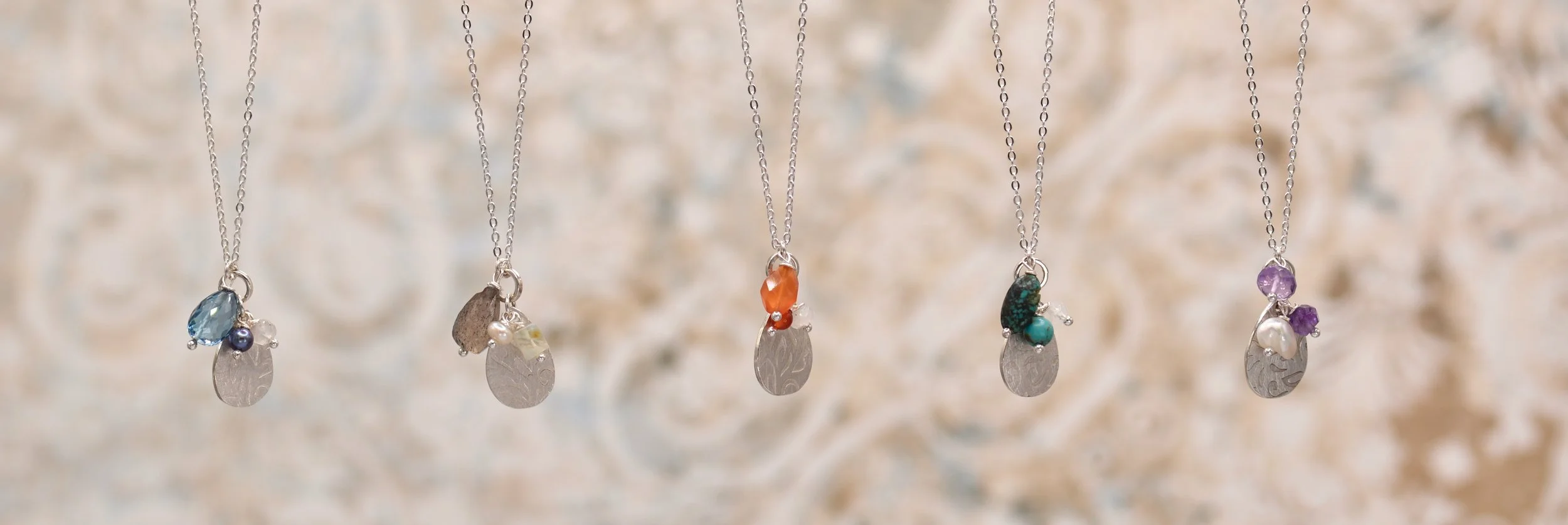 Five silver necklaces with various colored pendants and gemstones hanging vertically in front of a blurred ornate background.