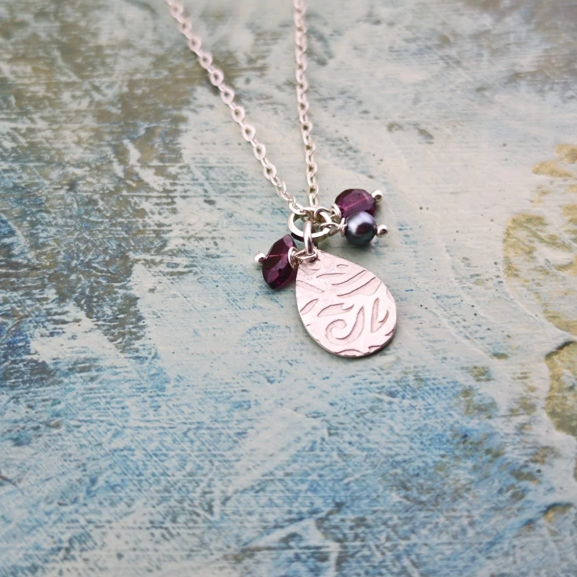 Silver necklace with a teardrop-shaped Silver pendant and multiple gemstones such as Garnet and and black pearl on a textured, painted surface.