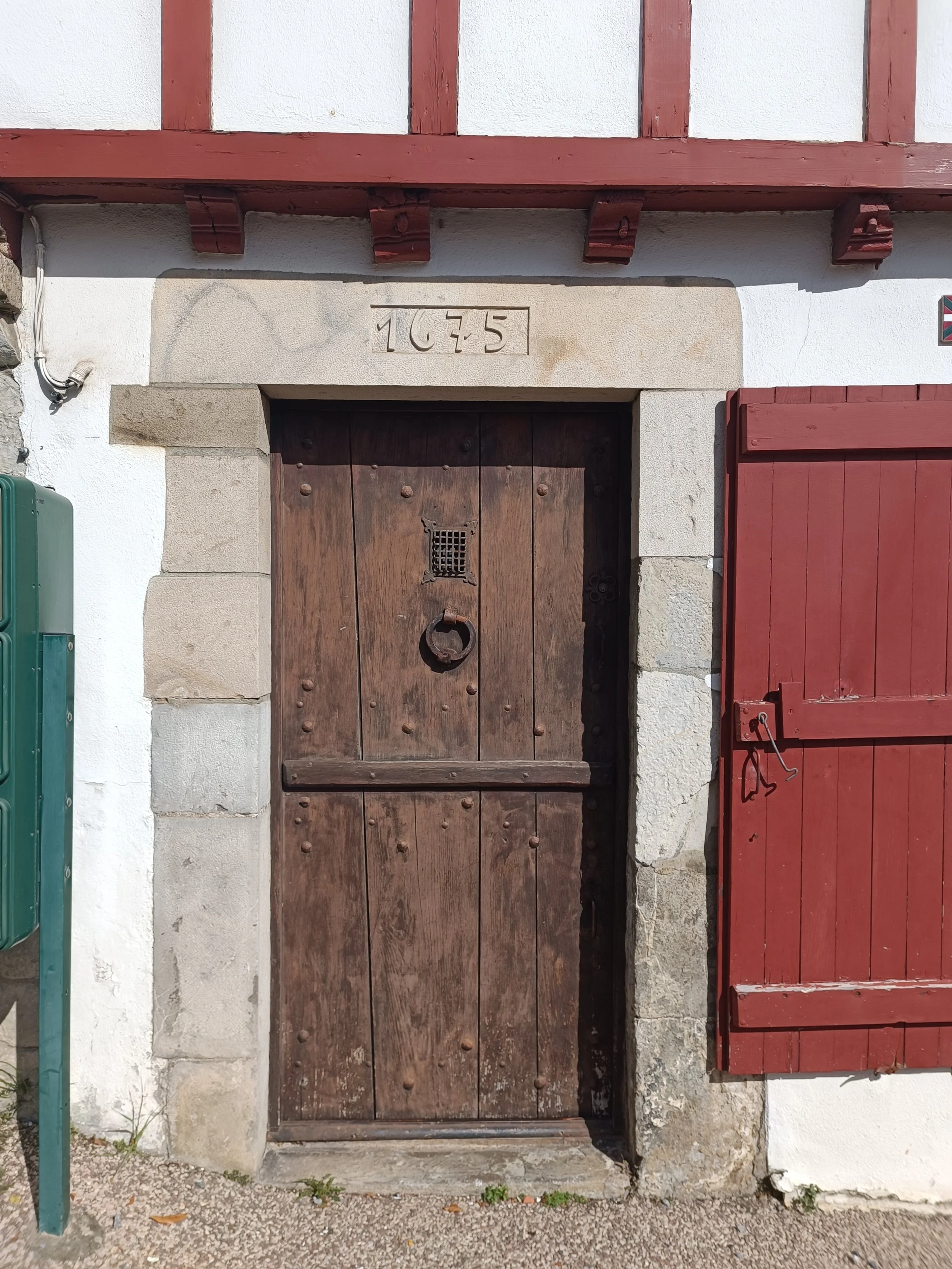 Wooden door in Urrugne, France with iron details, set in stone frame, with the year '1675' carved above, white building with red wooden accents and windows.