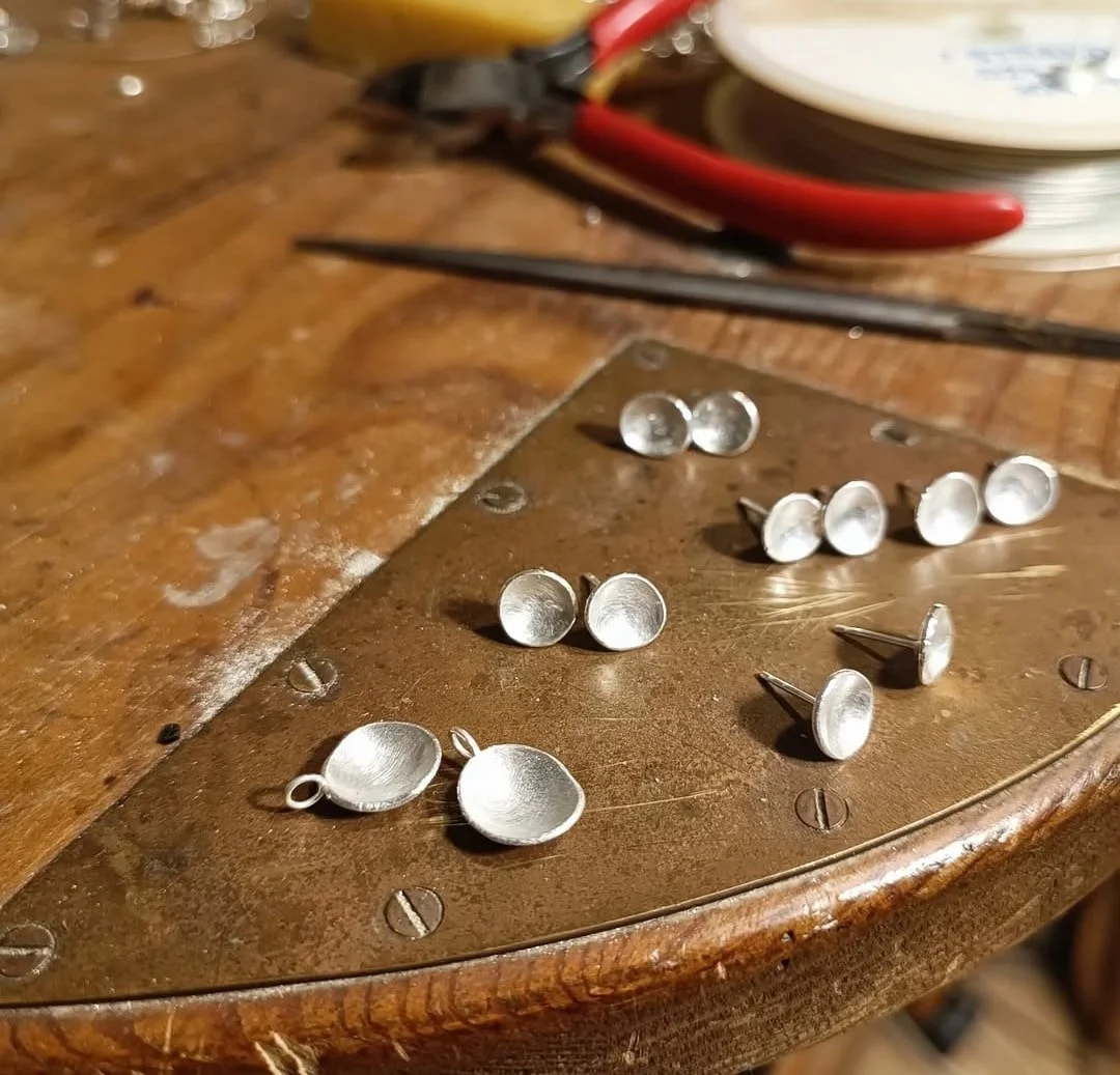 Small metal jewelry bezels with loops on a wooden workbench, jewelry-making tools and wires in the background.
