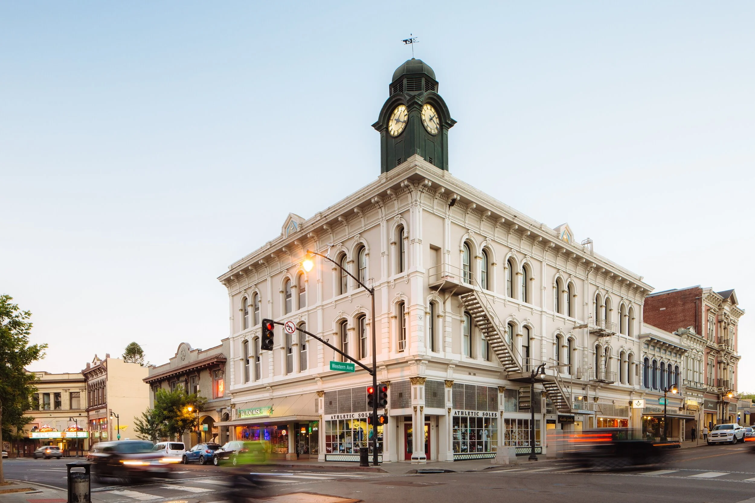 Historic white building with a clock tower at the corner of a busy street, with cars and pedestrians, and storefronts on the ground level.