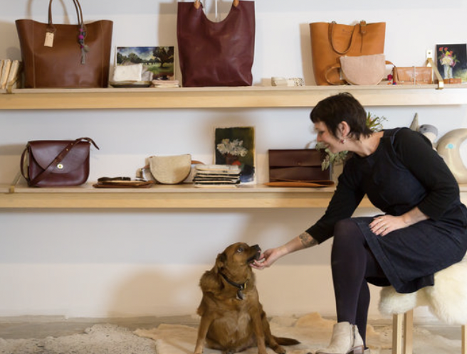 A woman sitting on a stool in a store, feeding a small brown dog. The background shows shelves with handbags, wallets, and other accessories.
