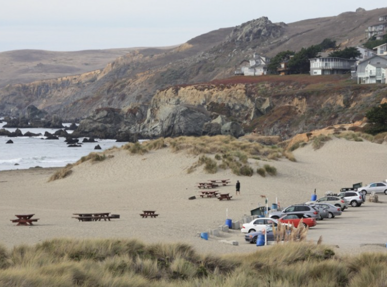 Beach with sand dunes, scattered picnic tables, parked cars, a person walking, rocky cliffs in the background, and ocean waves.