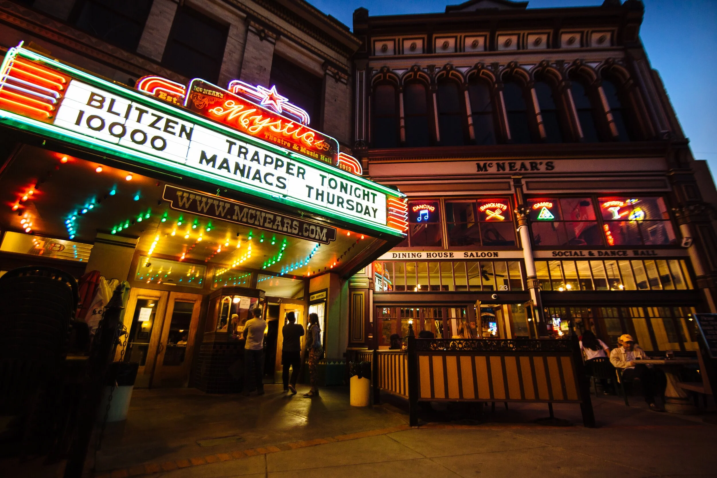 Night view of the McNears building with neon signs and an illuminated marquee advertising a show, dining, and dance hall. There are people standing outside and some seated at tables, enjoying the evening.