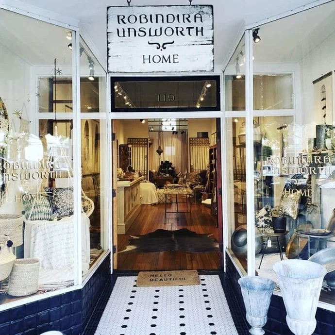 Storefront of Robindira Unsworth Home decor shop with large glass windows displaying interior furniture and decorations, entrance mat says "Hello Beautiful."