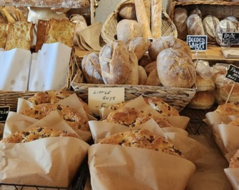 Baked bread and pastries displayed on a bakery counter with handwritten signs, including a basket of crusty bread loaves labeled 'Little Guys' and slices of pizza in parchment paper.
