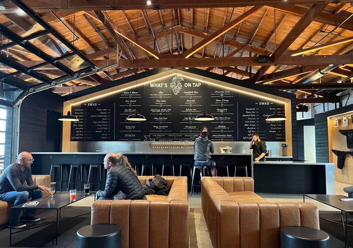 Inside a modern brewery or taproom with a high wooden ceiling, black walls, and a large chalkboard menu above the bar displaying various beer options. There are brown leather sofas and people sitting and chatting. Two staff members are behind the bar, one talking to a customer. The overall atmosphere is cozy and contemporary.