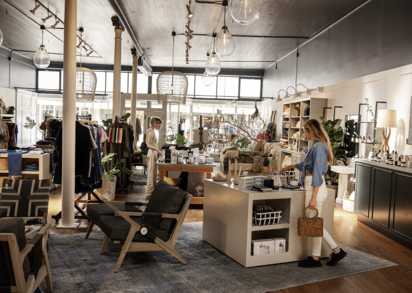 Interior of a boutique shop with clothing racks, a checkout counter, and decorative plants, with two women shopping and browsing.