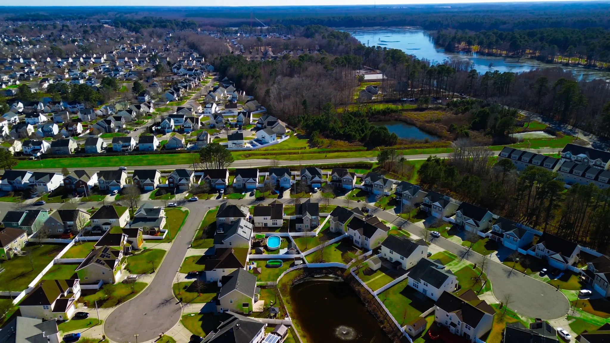 Aerial view of a suburban neighborhood with numerous houses, roads, green lawns, a small pond with fountain, and a nearby water body with boats and trees.