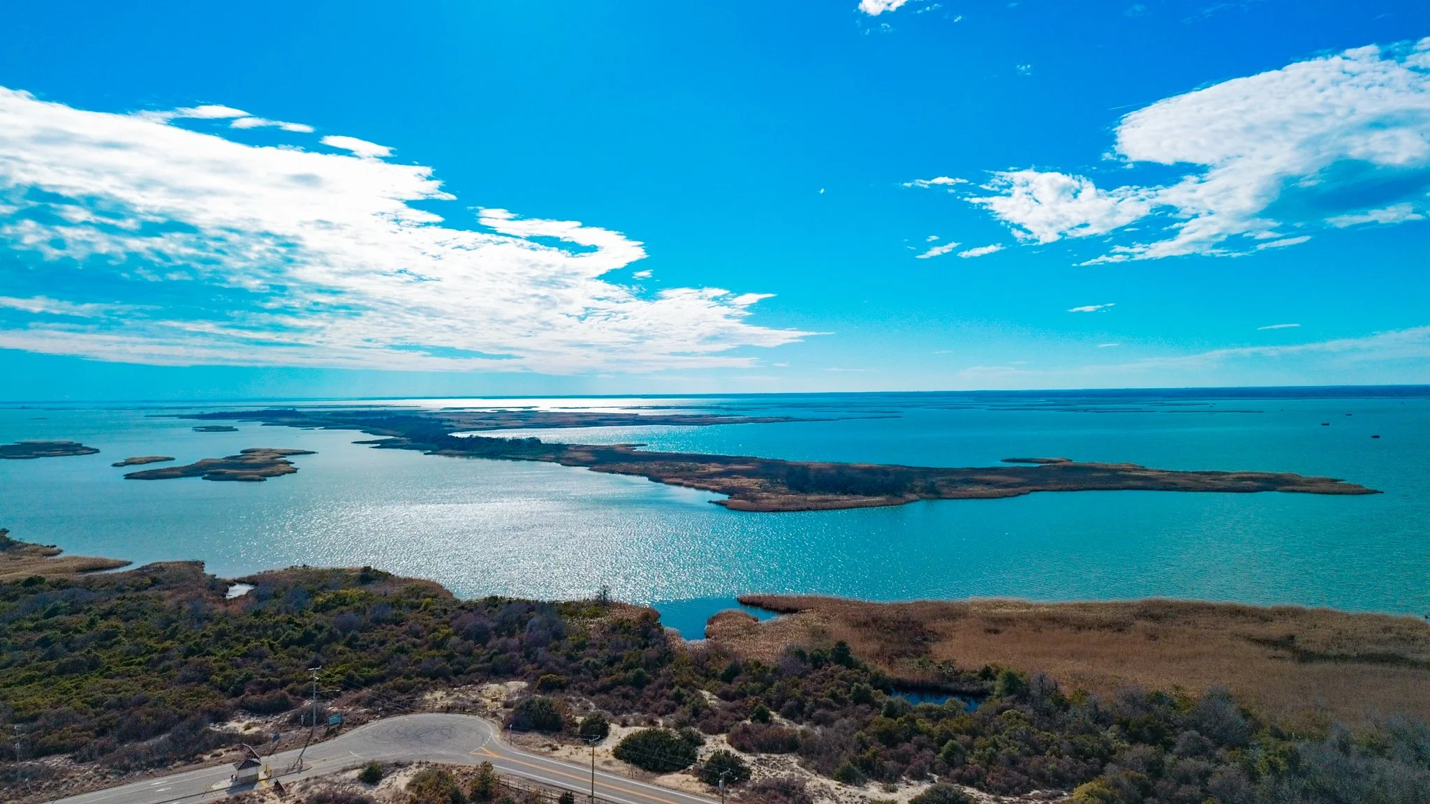 Aerial view of a large body of water with several small islands and patches of land, under a partly cloudy blue sky, with a road and vegetation in the foreground.